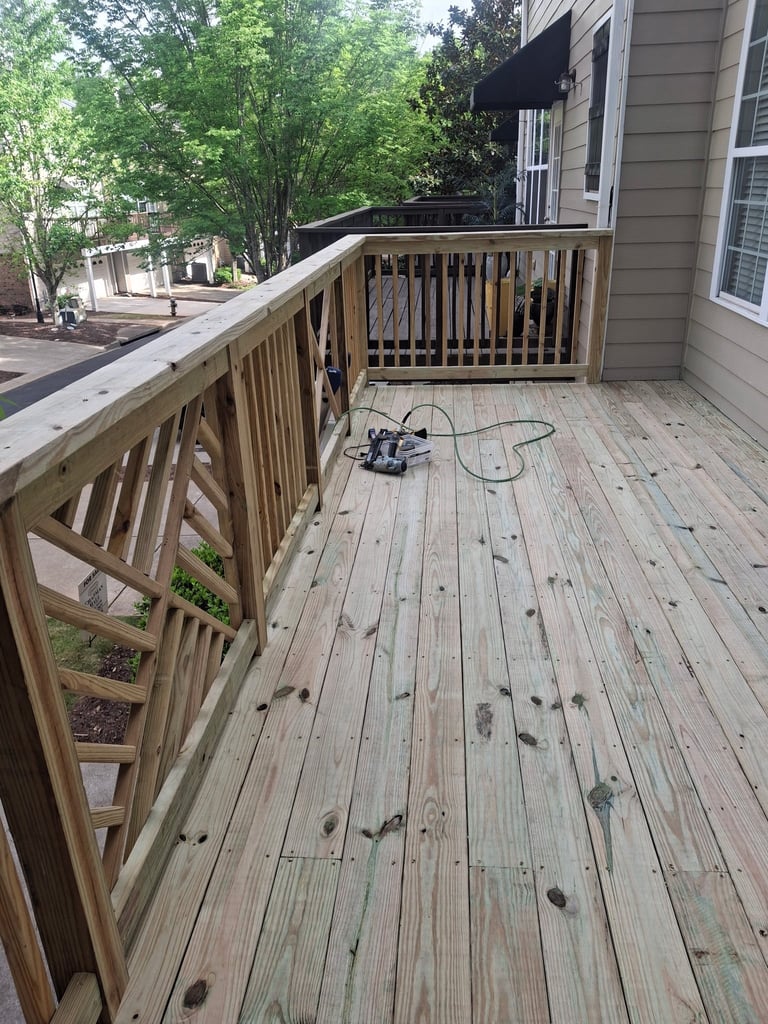 Wooden deck with railings attached to a house, featuring green trees in the background and a power tool on the weathered deck surface