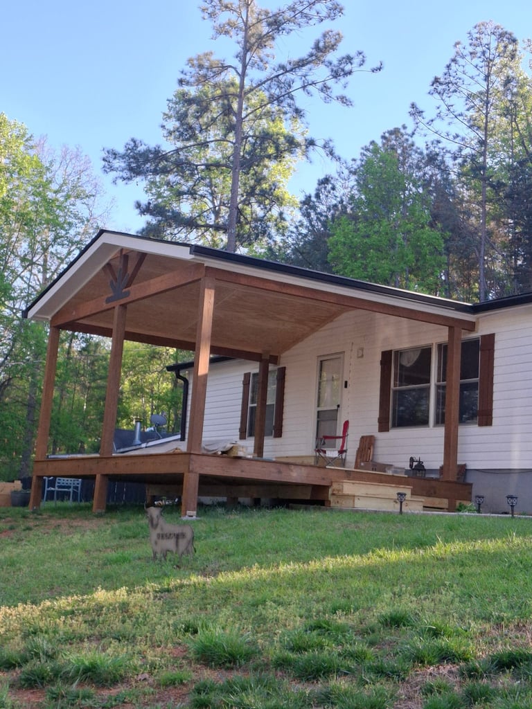 Small white mobile home with covered front porch surrounded by tall pine trees and green grass