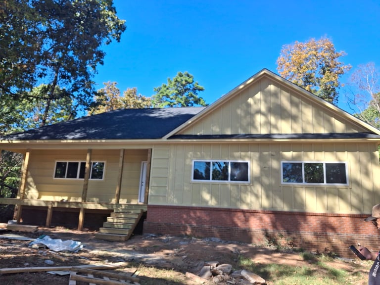 Single-story beige and brick residential house with dark roof and multiple windows surrounded by fall foliage and clear blue sky