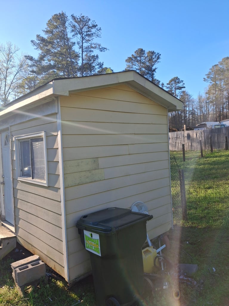 Tan colored garden shed with window and door, black trash bin beside it, wooden fence and trees in background