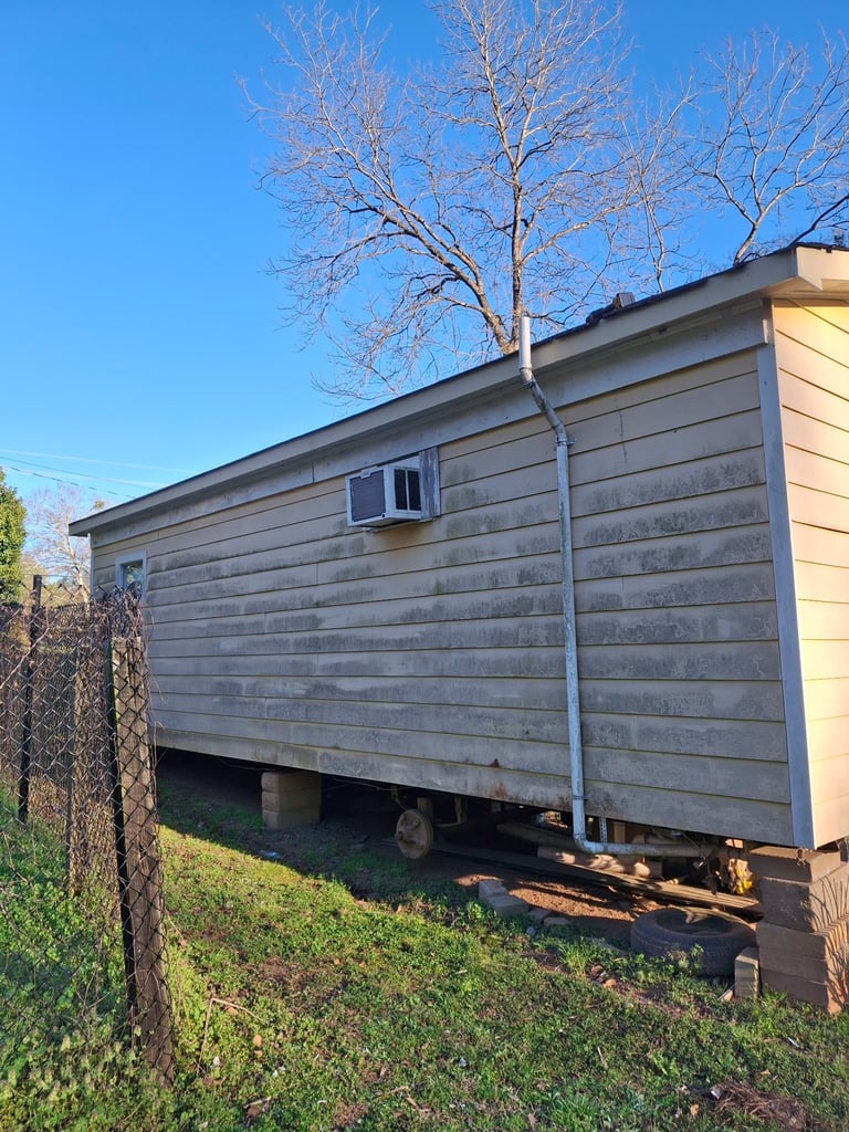 Mobile home elevated on blocks with bare tree, window unit AC, and chain-link fence against blue sky