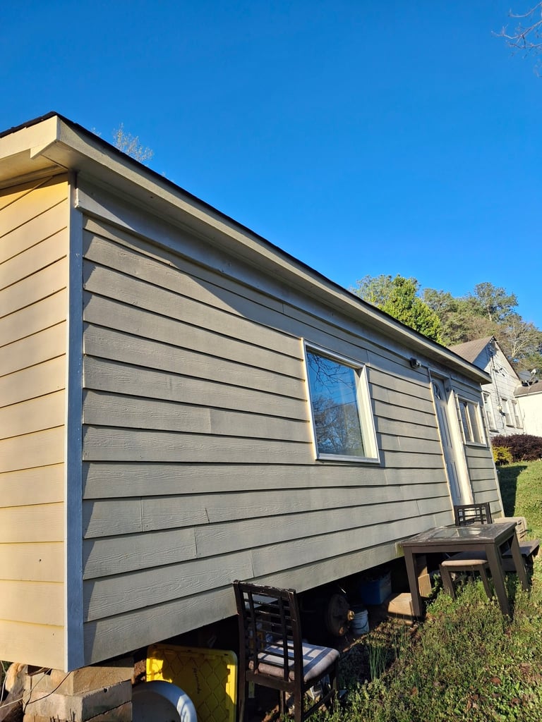 Mobile home or trailer with gray horizontal siding and windows, elevated on a foundation against a clear blue sky with trees in the background